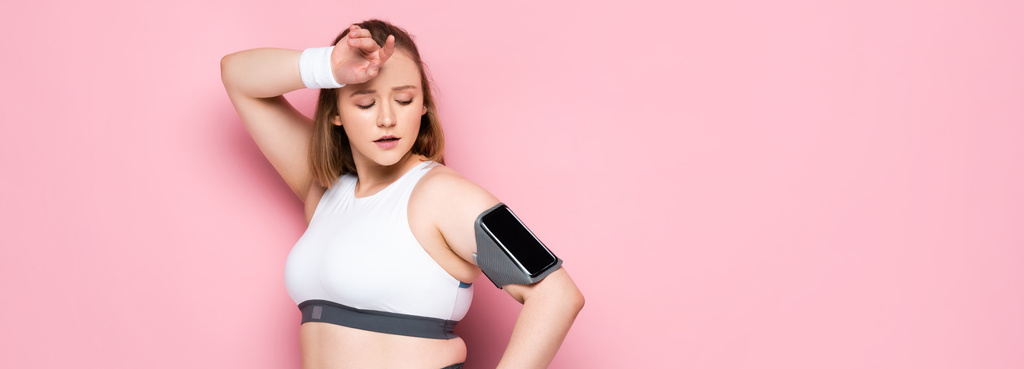 horizontaal beeld van moe overgewicht meisje kijken naar smartphone in armband terwijl het aanraken van het voorhoofd op roze - Foto, afbeelding