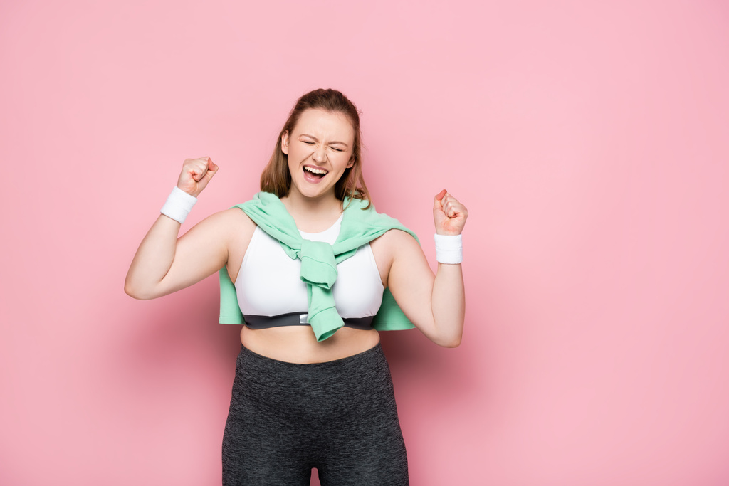 excitada chica con sobrepeso con sudadera sobre los hombros mostrando el gesto ganador con los ojos cerrados en rosa
 - Foto, imagen