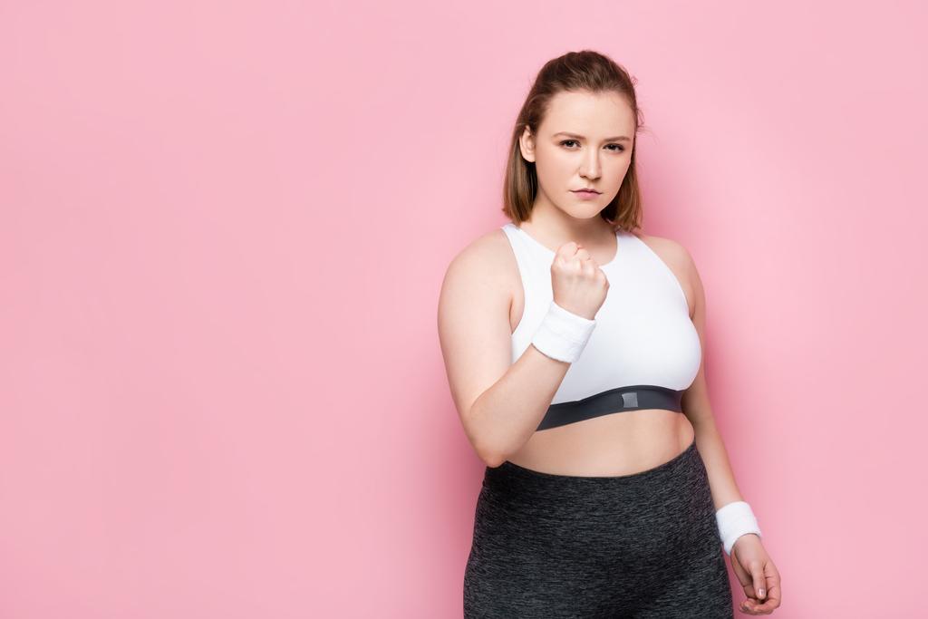 Wütendes übergewichtiges Mädchen zeigt geballte Faust und blickt in Kamera auf rosa - Foto, Bild