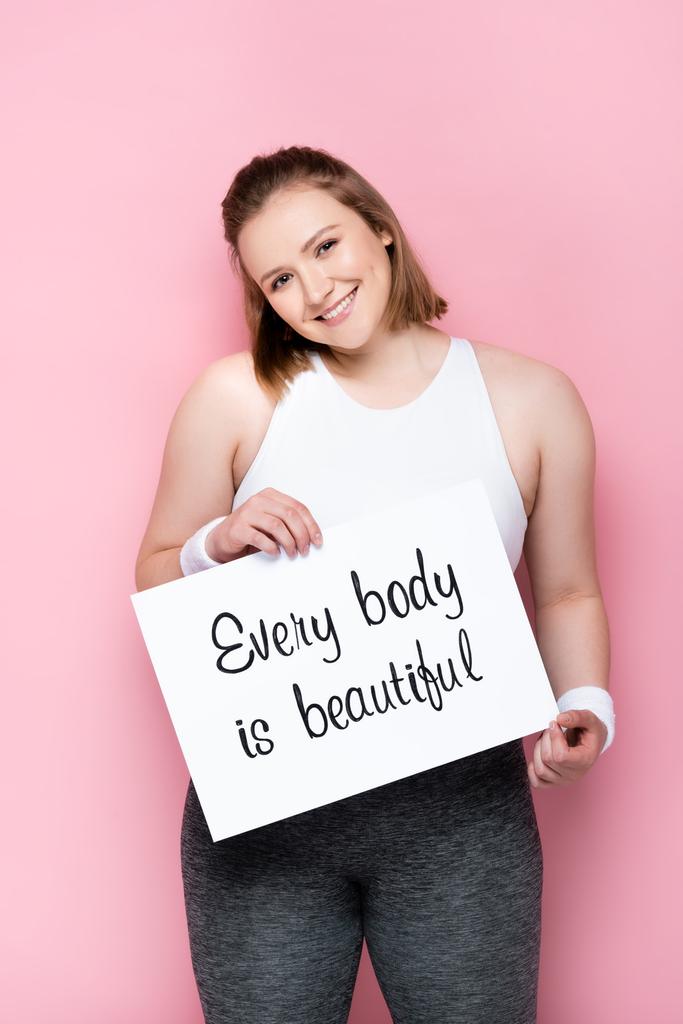 fröhliches übergewichtiges Mädchen hält Plakat mit jedem Körper ist schöne Inschrift auf rosa - Foto, Bild