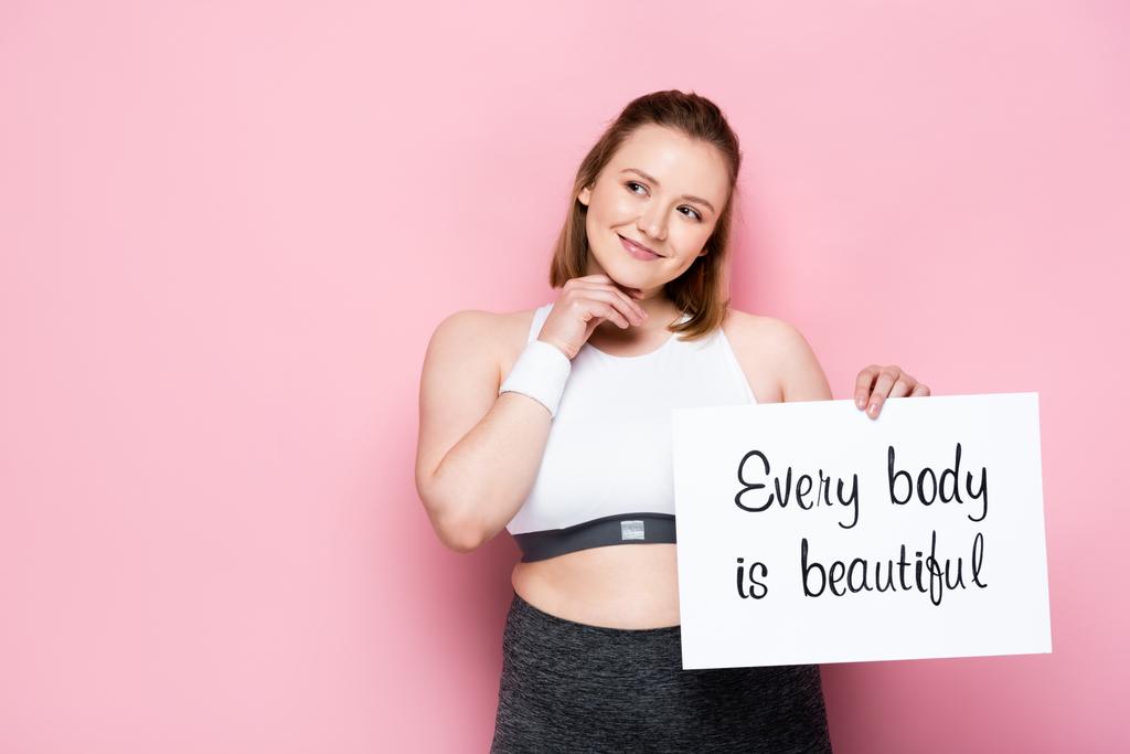 smiling overweight girl holding placard with every body is beautiful inscription on pink - Photo, Image