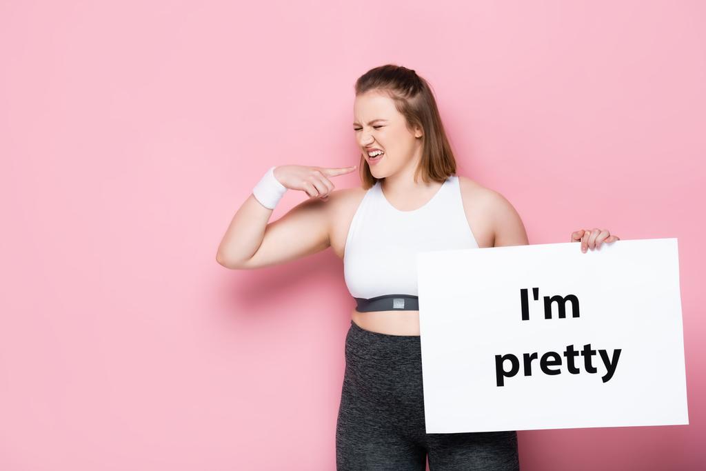 nespokojený nadváha dívka šklebí při držení plakátu s jsem pěkný nápis na růžové  - Fotografie, Obrázek