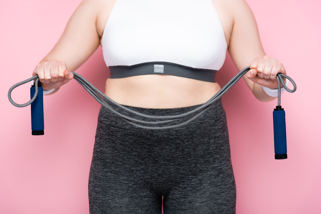 vista parziale della ragazza in sovrappeso in abbigliamento sportivo che tiene la corda da salto sul rosa - Foto, immagini