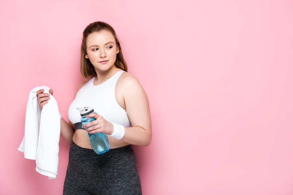 zelfverzekerde overgewicht meisje met sport fles en witte handdoek terwijl weg te kijken op roze - Foto, afbeelding