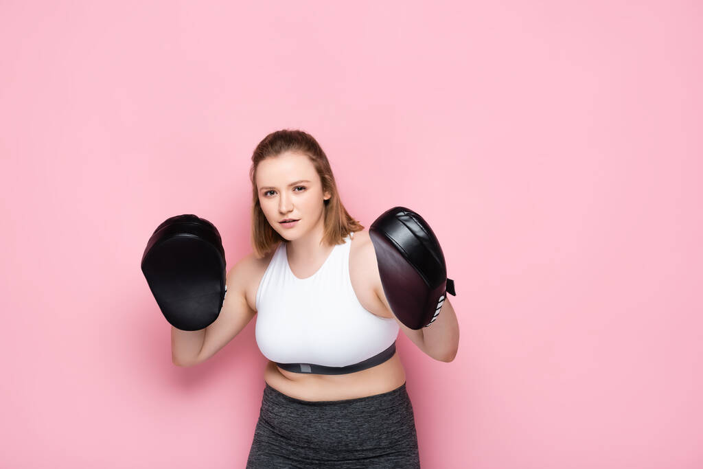 συμπυκνωμένο υπέρβαρο κορίτσι άσκηση με μαξιλάρια πυγμαχίας, ενώ κοιτάζοντας κάμερα σε ροζ - Φωτογραφία, εικόνα