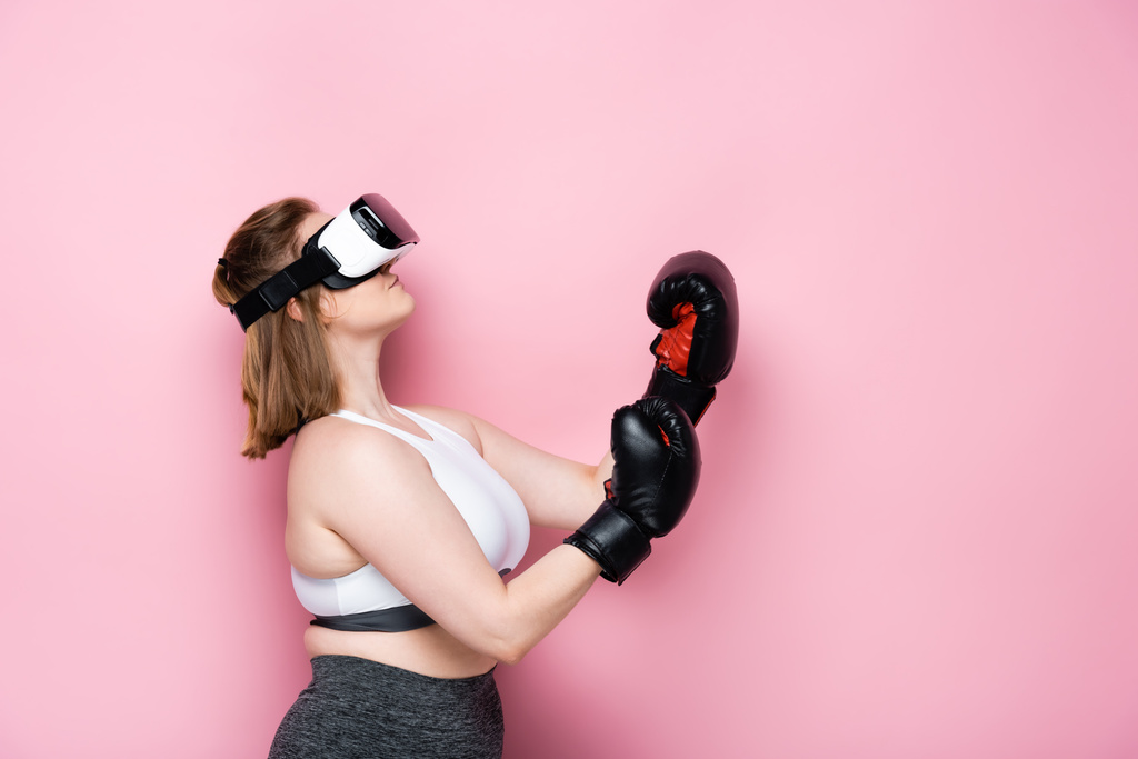 vista lateral de la chica con sobrepeso en auriculares vr y guantes de boxeo mirando hacia arriba en rosa
 - Foto, imagen