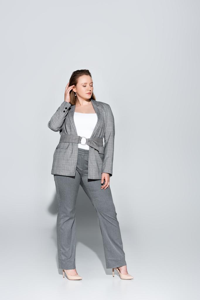 full length view of stylish overweight girl touching hair while posing with closed eyes on grey - Photo, Image