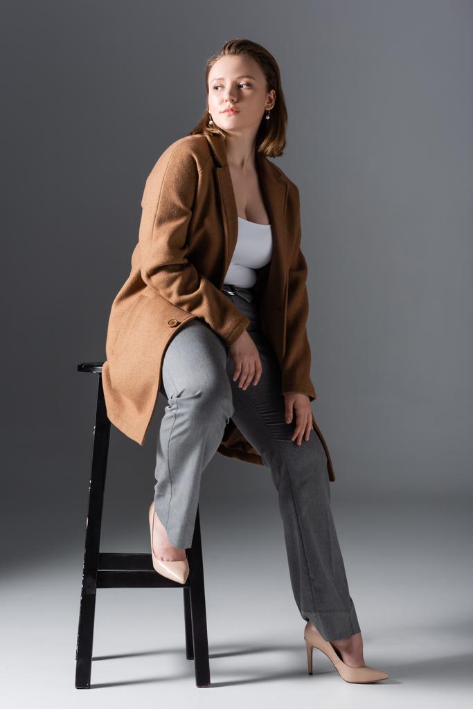 volledige weergave van zelfverzekerde, stijlvolle overgewicht meisje poseren op hoge stoel en weg te kijken op grijs - Foto, afbeelding