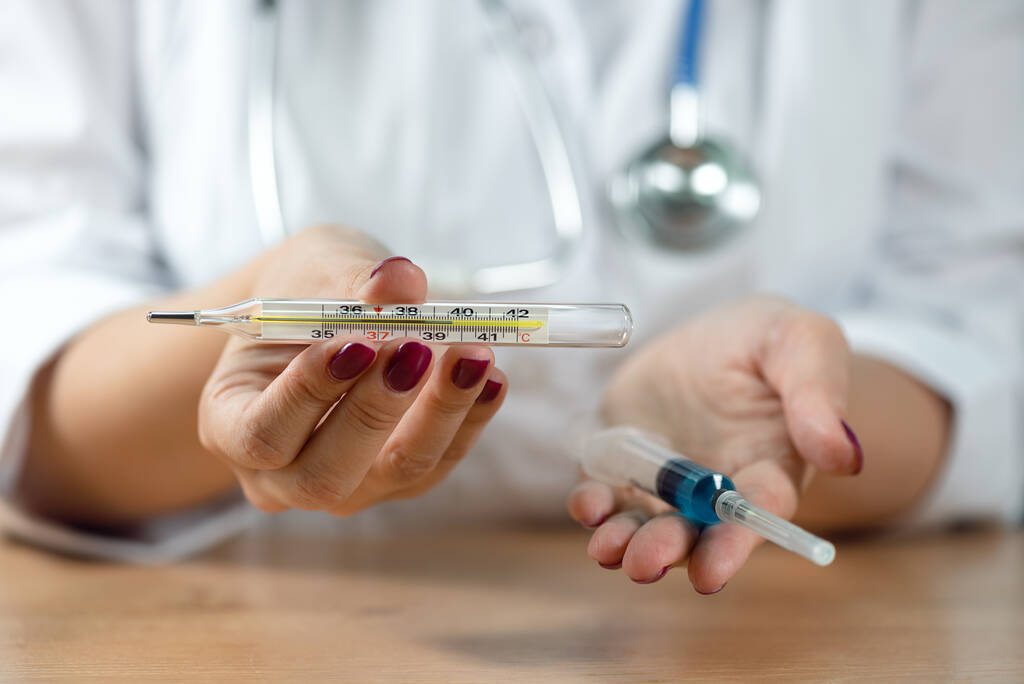 Szczepienie przeciw grypie. Lekarz kobieta trzyma w rękach termometr z wysoką temperaturą i strzykawkę ze szczepionką. - Zdjęcie, obraz