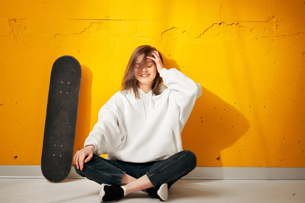 Ένα νεαρό χαρούμενο χαμογελαστό κορίτσι με λευκή κουκούλα με σκέιτμπορντ ποζάρει σε έναν κίτρινο τσιμεντένιο τοίχο.. - Φωτογραφία, εικόνα