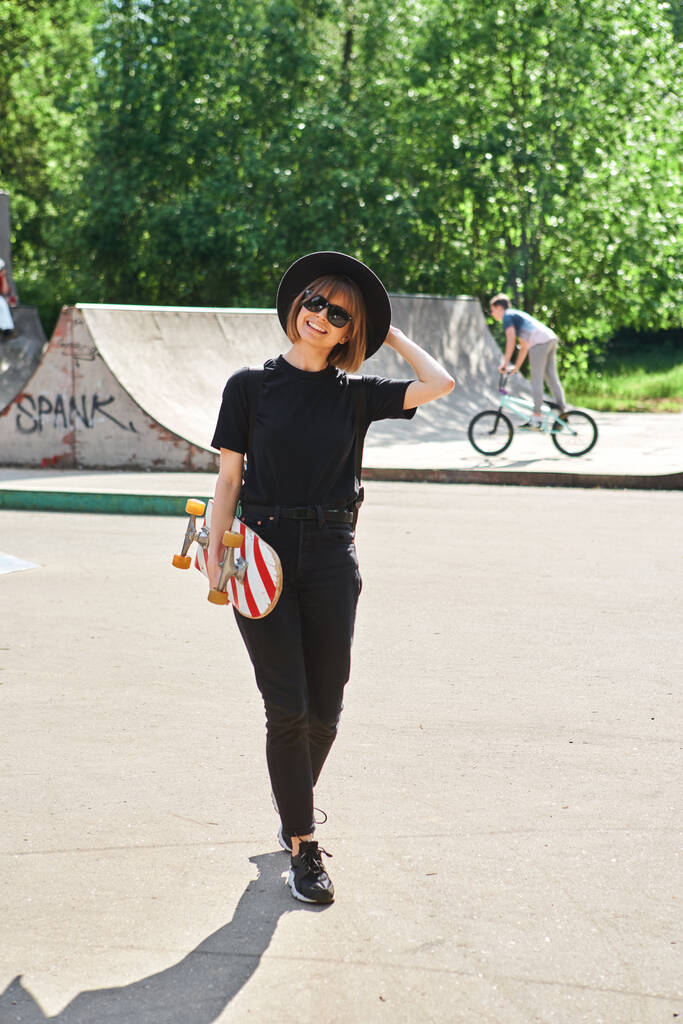 Pěkně usměvavá dospívající dívka nesoucí skateboard procházky ve skateparku ve slunečních brýlích, klobouku a černém oblečení - Fotografie, Obrázek