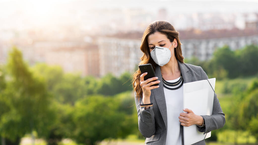 Junge Geschäftsfrau mit Mundschutz draußen während der Covid-19-Coronavirus-Gesundheitskrise. Professionelle Frau benutzt ihr Smartphone. - Foto, Bild