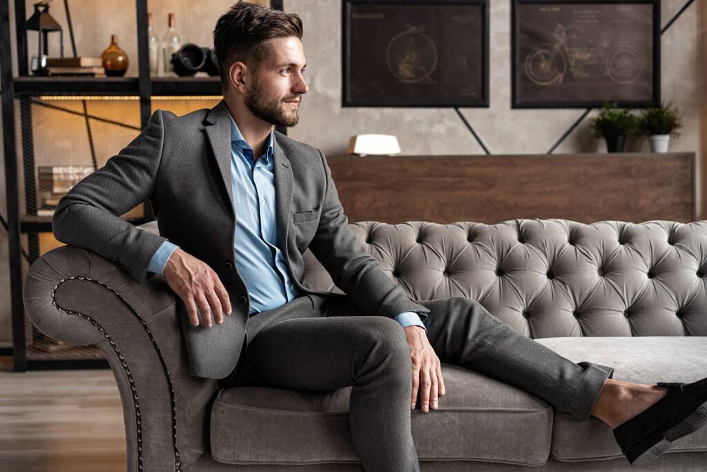 Cool und gutaussehend. Gut aussehender junger Mann in vollem Anzug sitzt auf dem Sofa, während er Zeit drinnen verbringt. - Foto, Bild