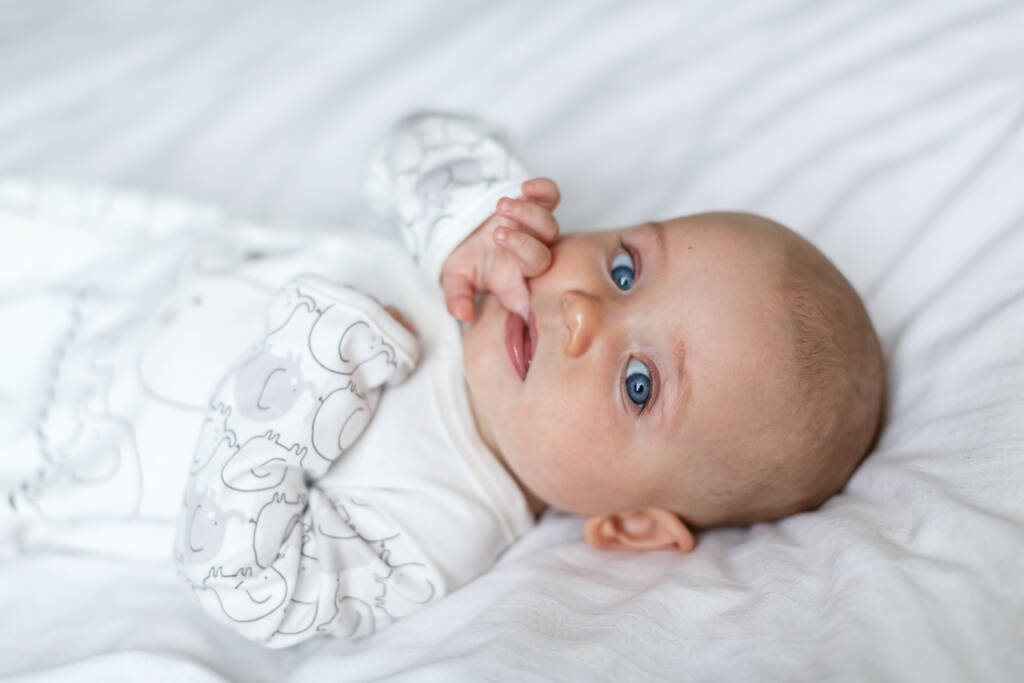 小さい赤ん坊は親の寝室の白いシートのベッドの上に横になって笑っている。新生児の白いジャンプスーツ。かわいい赤ん坊がベッドの上に横たわっていた。カメラを見る新生児のクローズアップポートレート - 写真・画像
