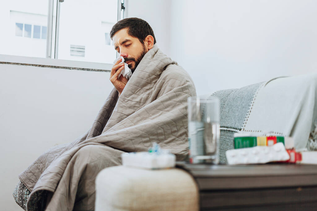 Mann, der sich zu Hause auf der Couch mit Erkältungs- und Grippesymptomen krank fühlt. Medizin und Glas Wasser im Vordergrund. - Foto, Bild