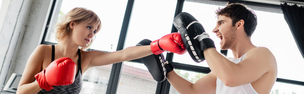 horizontaal beeld van sportief meisje in bokshandschoenen ponsen man in boksen pads  - Foto, afbeelding