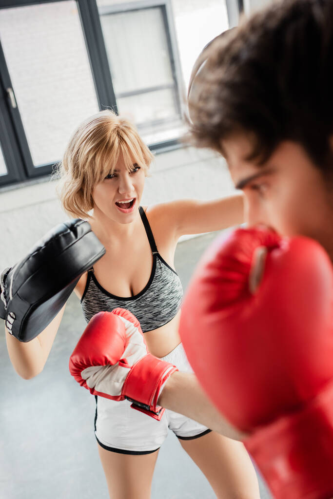 ジムでスポーツマンと運動するボクシングパッドの感情的な女の子の選択的な焦点  - 写真・画像