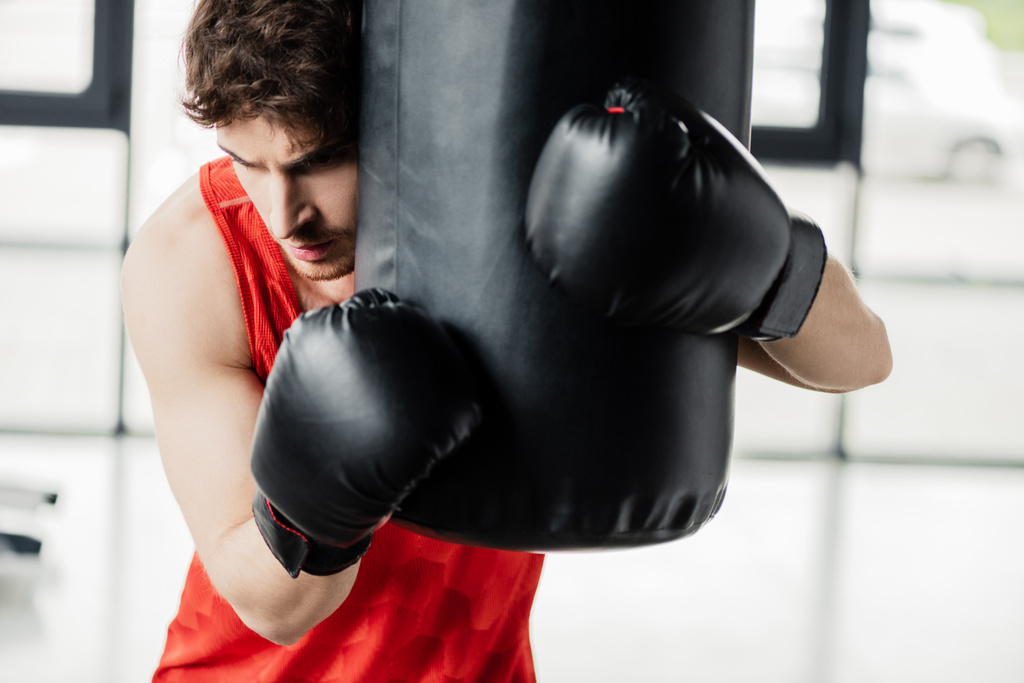 Müder Mann in Sportbekleidung und schwarzen Boxhandschuhen berührt Boxsack in Turnhalle  - Foto, Bild