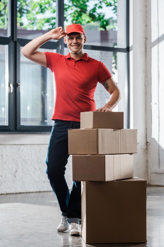 χαρούμενος και τατουάζ delivery man αγγίζοντας το καπάκι, ενώ στέκεται κοντά σε κουτιά χαρτοκιβωτίων  - Φωτογραφία, εικόνα