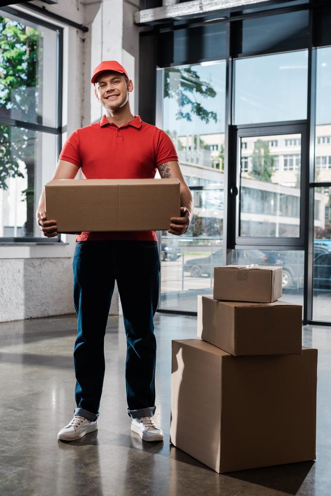 cheerful delivery man in cap holding carton box and looking at camera - Photo, Image