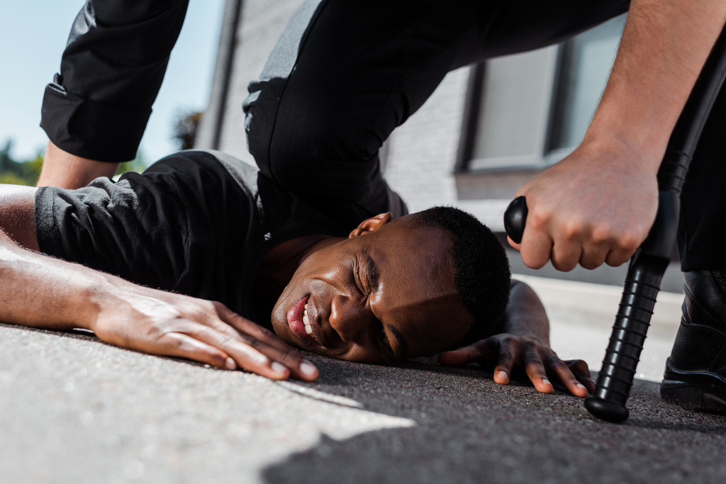 επιλεκτική εστίαση του κρατούμενου και τραυματισμένου Αφροαμερικανού άνδρα που κείτεται στο έδαφος κοντά σε αστυνομικό με ρόπαλο, έννοια του ρατσισμού  - Φωτογραφία, εικόνα