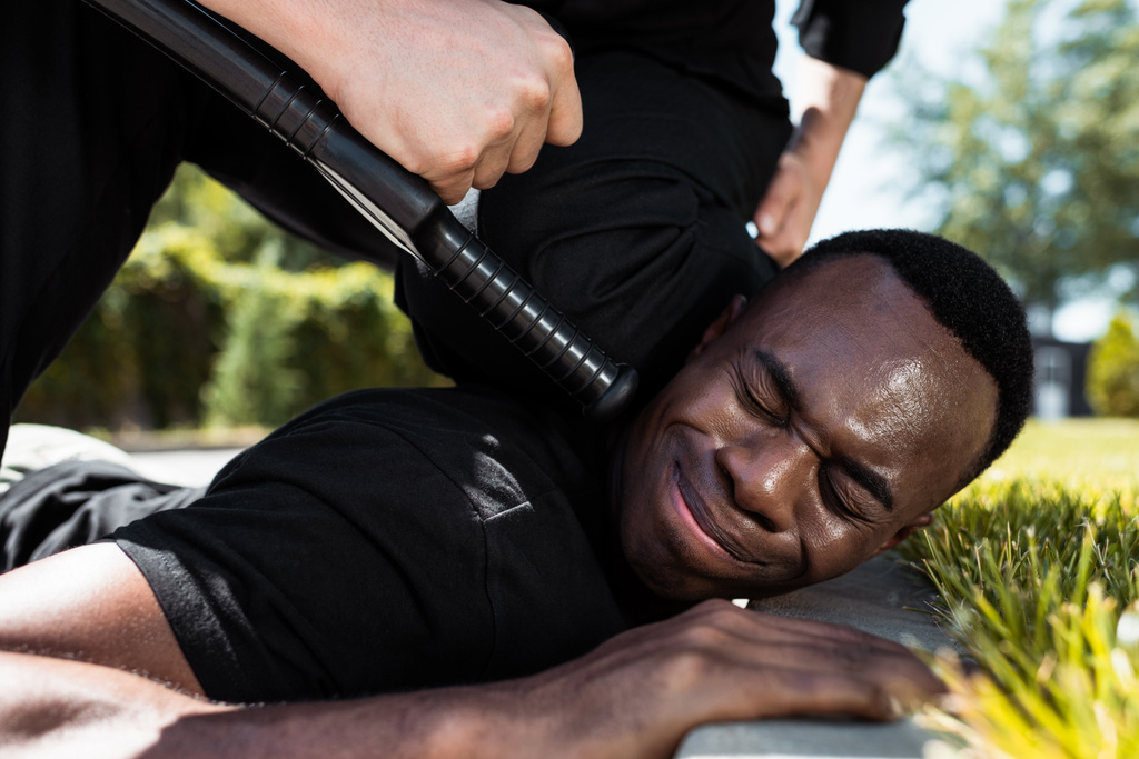 selective focus of detained african american man suffering from pain while lying near policeman with baton, racism concept  - Photo, Image
