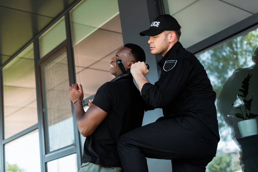 policía con gorra sosteniendo bastón policial cerca de la garganta del hombre afroamericano esposado, concepto de racismo
  - Foto, Imagen