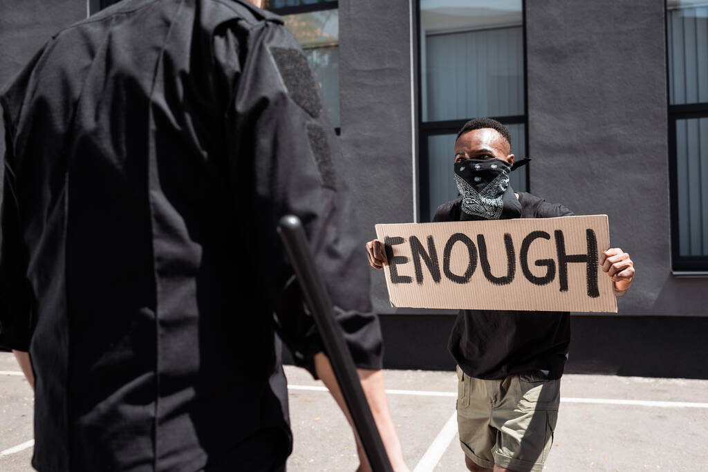 enfoque selectivo del hombre afroamericano con bufanda en la cara con pancarta con suficientes letras cerca de policía con bastón en la calle, concepto de racismo
  - Foto, Imagen