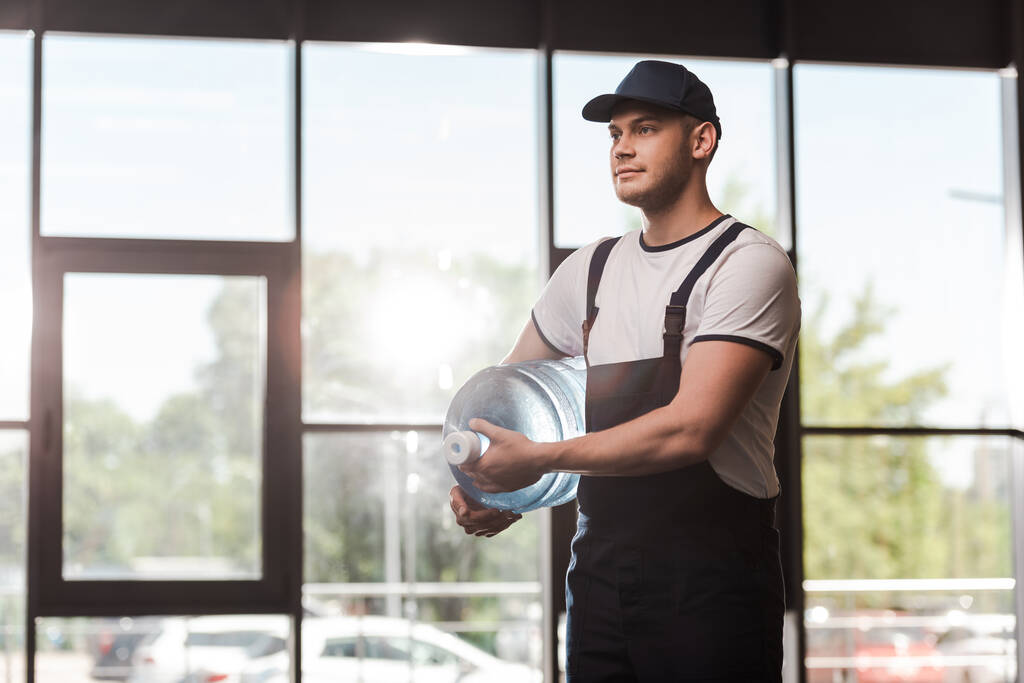 handsome delivery man in uniform and cap holding bottled water  - Photo, Image