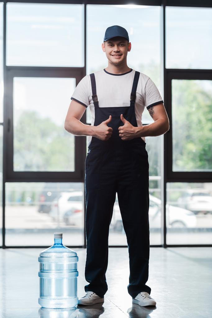 allegro uomo di consegna in uniforme mostrando pollici verso l'alto vicino gallone di acqua in bottiglia  - Foto, immagini