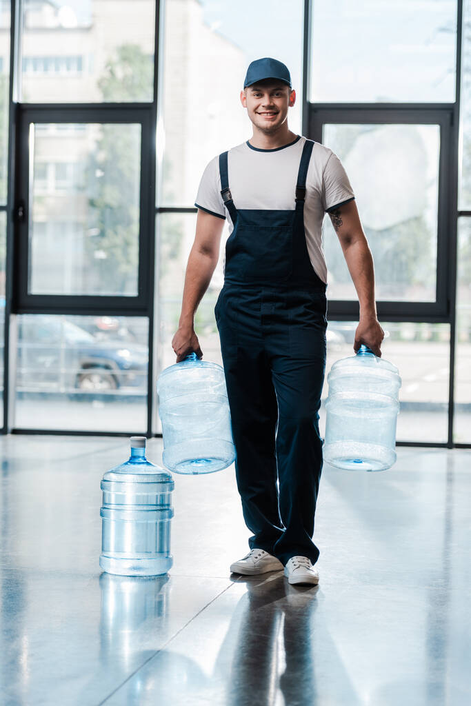 Vidám futár egyenruhában, üres palackokkal a kezében, közel egy gallon vízhez.  - Fotó, kép