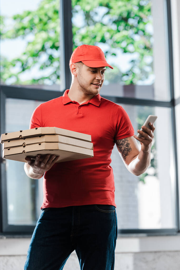 toimitusmies korkissa katsomassa älypuhelinta ja pitämällä pizzalaatikoita  - Valokuva, kuva