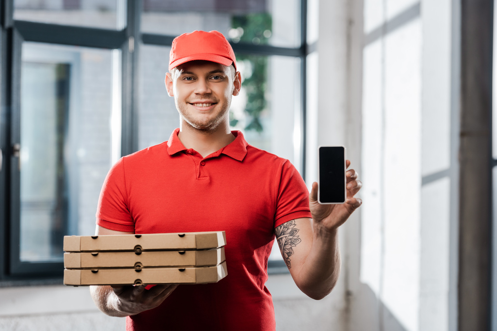 happy delivery man in cap holding smartphone met blanco scherm en pizza dozen  - Foto, afbeelding