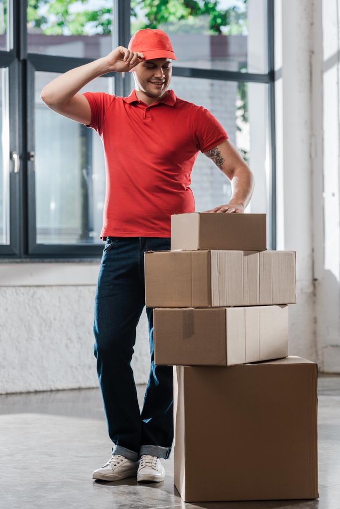 cheerful delivery man touching cap and looking at carton boxes  - Photo, Image