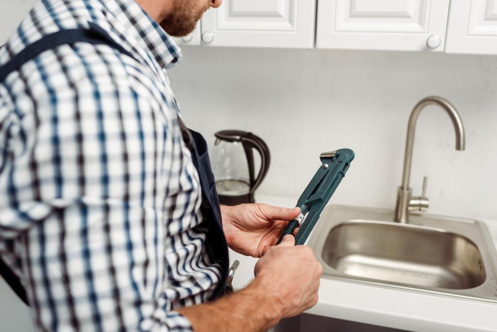 Zugeschnittene Ansicht des Klempners, der Schraubenschlüssel in der Nähe von Wasserhahn und Spüle in der Küche hält  - Foto, Bild