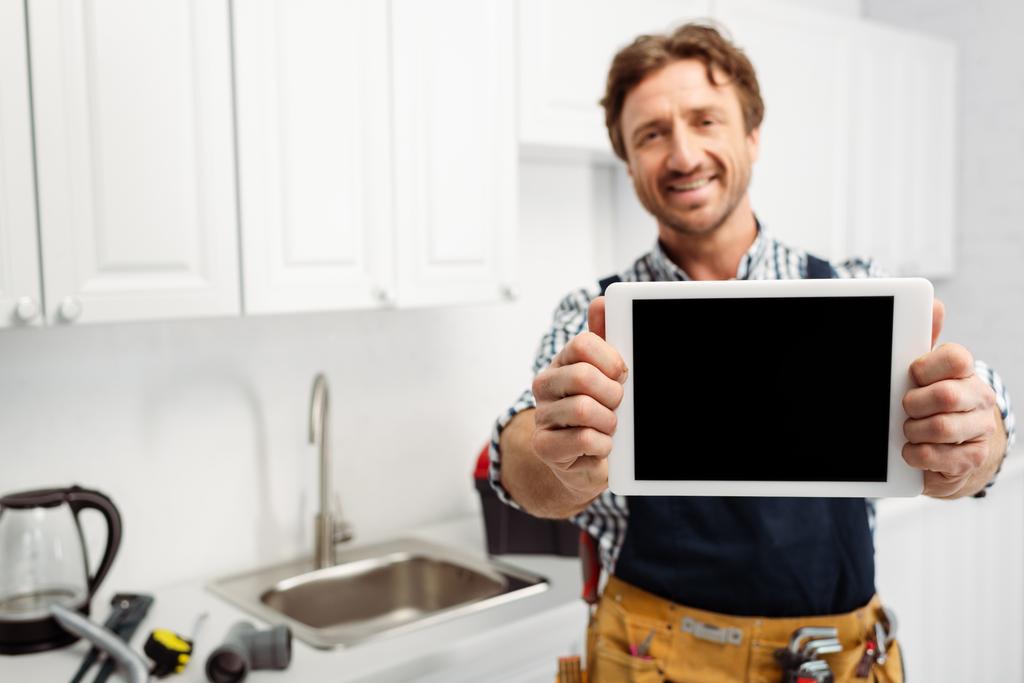 Selective focus of smiling plumber showing digital tablet with blank screen in kitchen  - Photo, Image