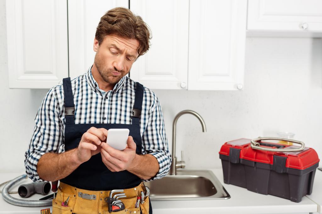 Encanador pensivo usando smartphone perto de caixa de ferramentas e pia na cozinha
  - Foto, Imagem