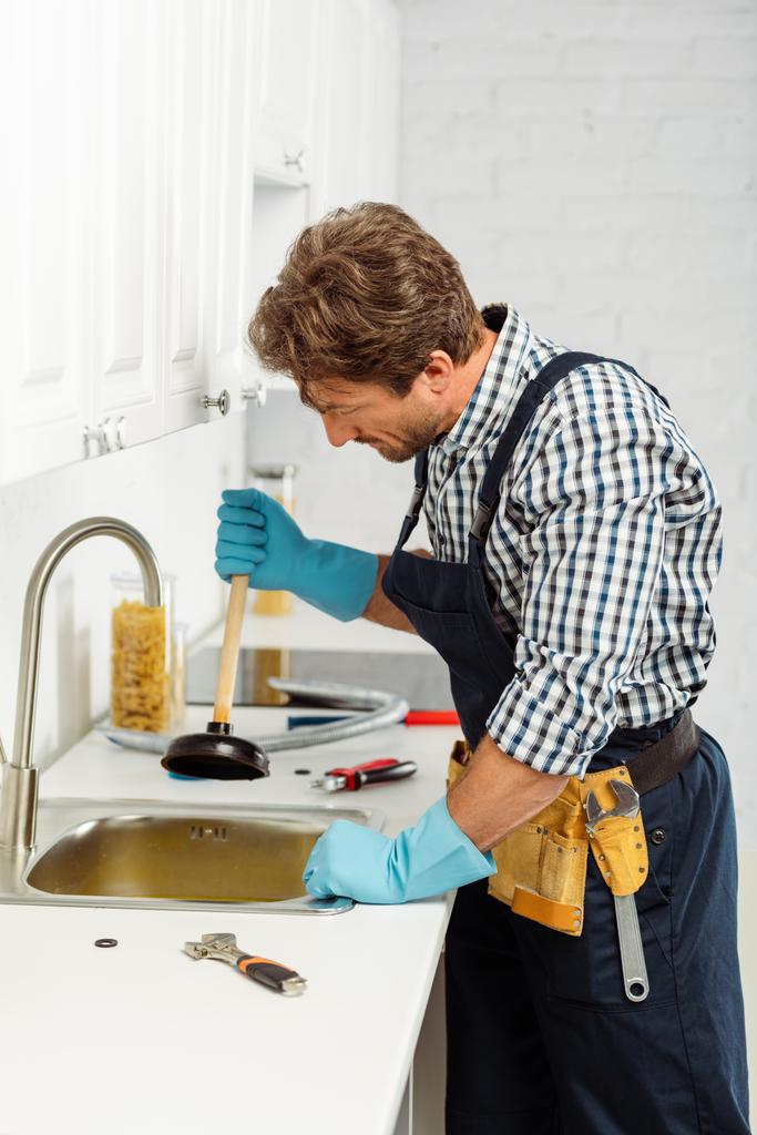 Boční pohled instalatéra v gumových rukavicích držící píst při upevňování kuchyňského dřezu  - Fotografie, Obrázek