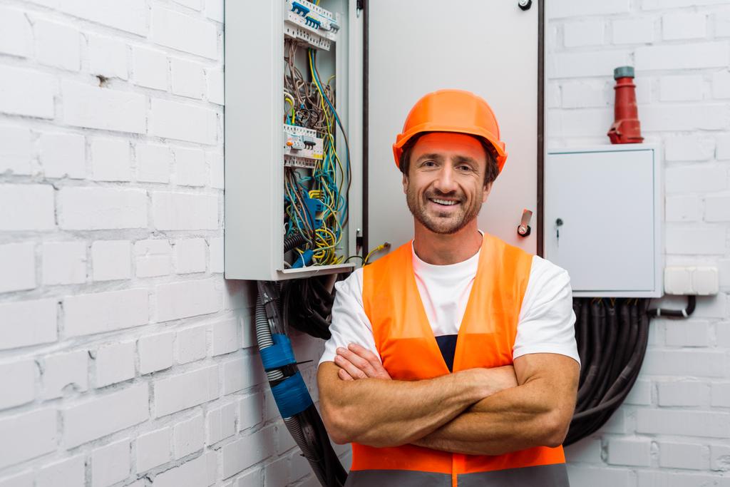 Handsome electrician with crossed arms smiling at camera near electric panel - Photo, Image