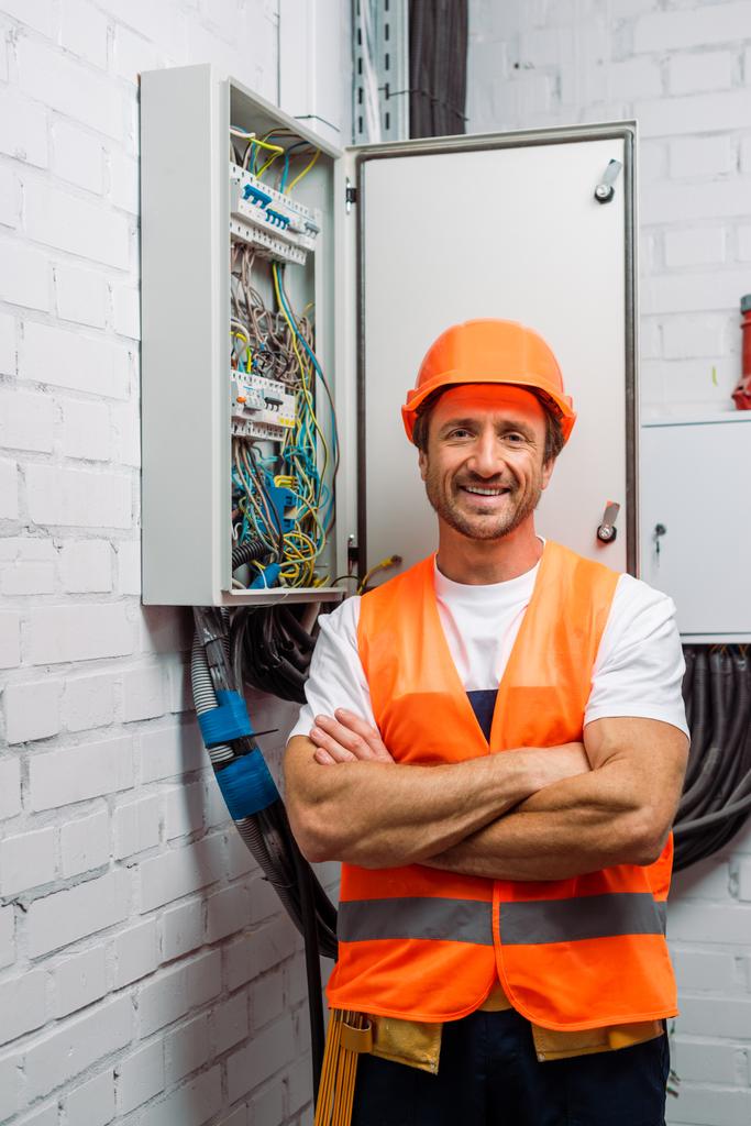Eletricista bonito em chapéu duro e colete de segurança sorrindo para a câmera perto da caixa de distribuição elétrica
 - Foto, Imagem
