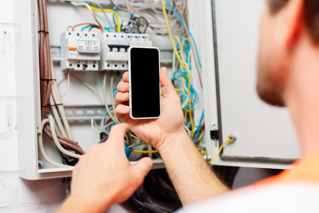 Selektiver Fokus des Elektrikers, der sein Smartphone in der Nähe eines Stromverteilerkastens hält - Foto, Bild