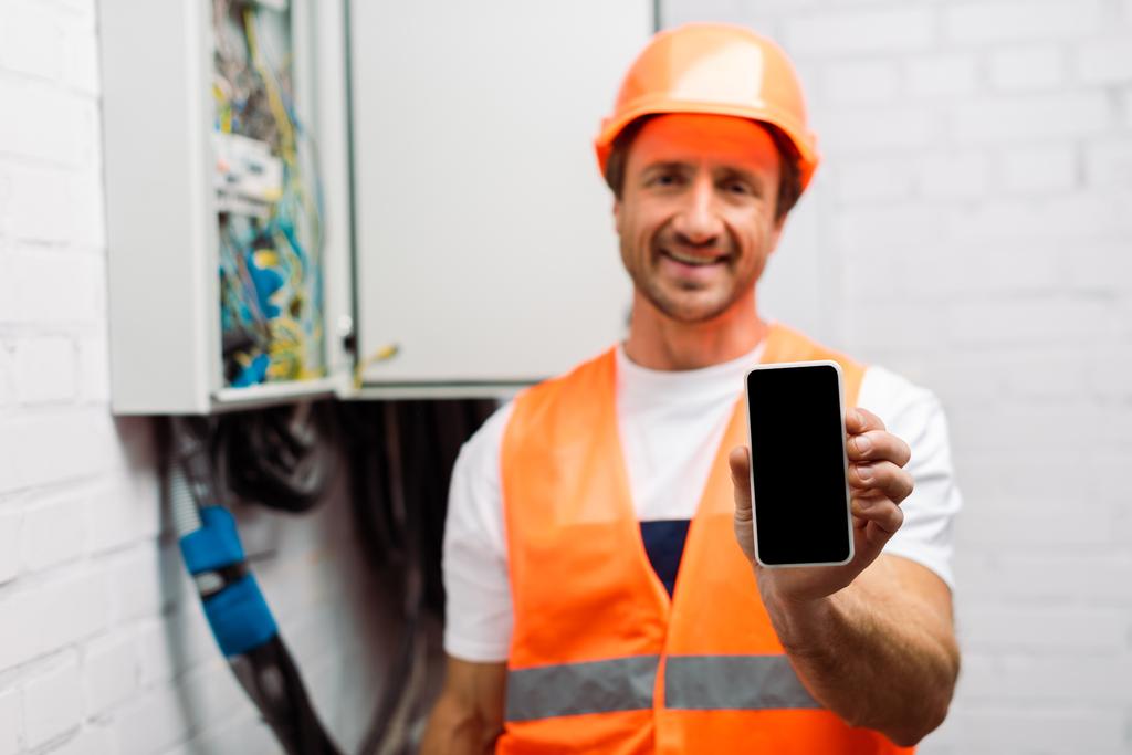 Selektiver Fokus eines lächelnden Elektrikers, der ein Smartphone mit leerem Bildschirm in der Nähe eines elektrischen Verteilerkastens hält - Foto, Bild