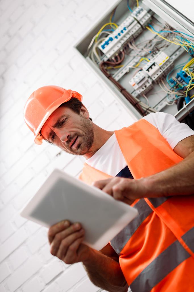 Selective focus of handsome workman in safety vest and helmet using digital tablet near electric box  - Photo, Image