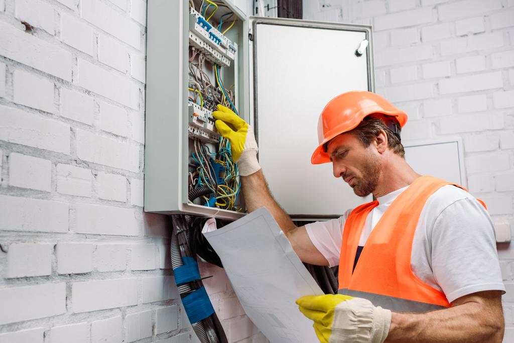 Schöner Elektriker in Handschuhen schaut sich den Bauplan an und repariert Stromverteilerkasten - Foto, Bild