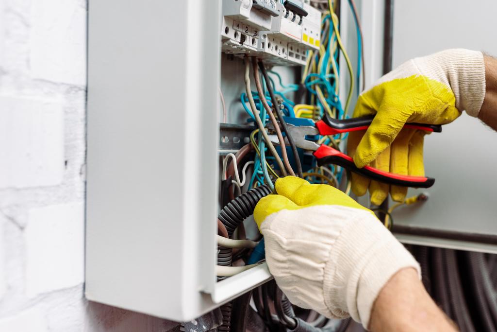 Vista recortada del trabajador con guantes sujetando alicates mientras fija la caja de distribución eléctrica
 - Foto, Imagen