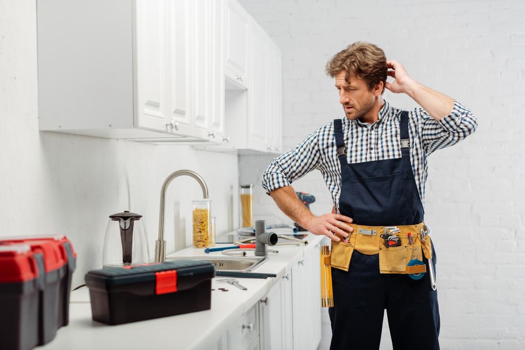 Выборочный фокус вдумчивого сантехника, смотрящего на кухонную раковину рядом с инструментами и ящиками для инструментов на кухонном столе
  - Фото, изображение