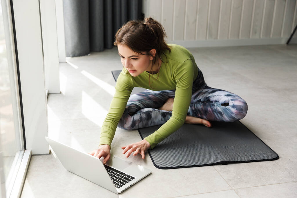 Foto de la deportista morena enfocada usando el ordenador portátil mientras hace ejercicio en la estera en la habitación acogedora
 - Foto, Imagen