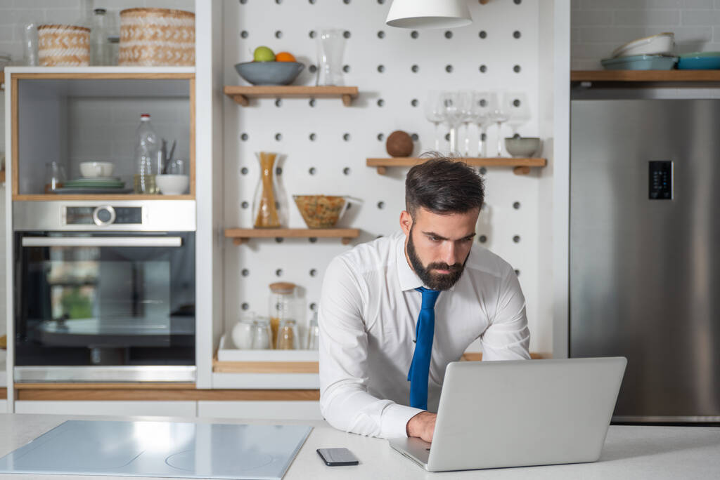 Młody brodaty biznesmen stojący w kuchni i sprawdzający firmową listę pracy pod kątem nowego biznesplanu i projektu na laptopie - Zdjęcie, obraz