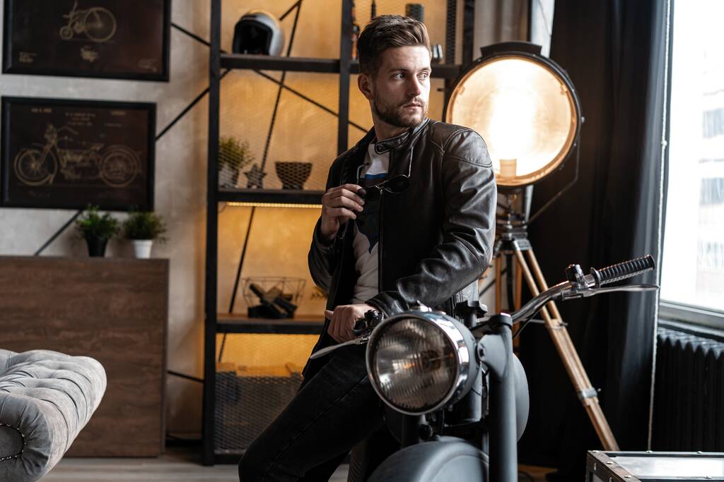 Стильный молодой человек в кожаной куртке сидит на мотоцикле
. - Фото, изображение