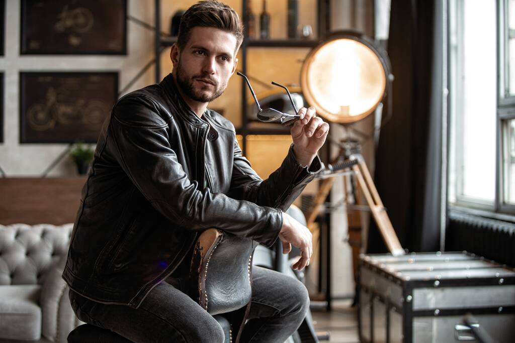 Handsome young man in leather jacket looking away while sitting on the stool - Photo, Image